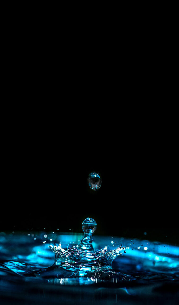 Ein illuminierter Wassertropfen, welcher nach oben sprizt, vor schwarzem Hintergrund.